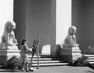Black and white photograph of a 1940s college student painting on a easel next to two stone Fu Dogs